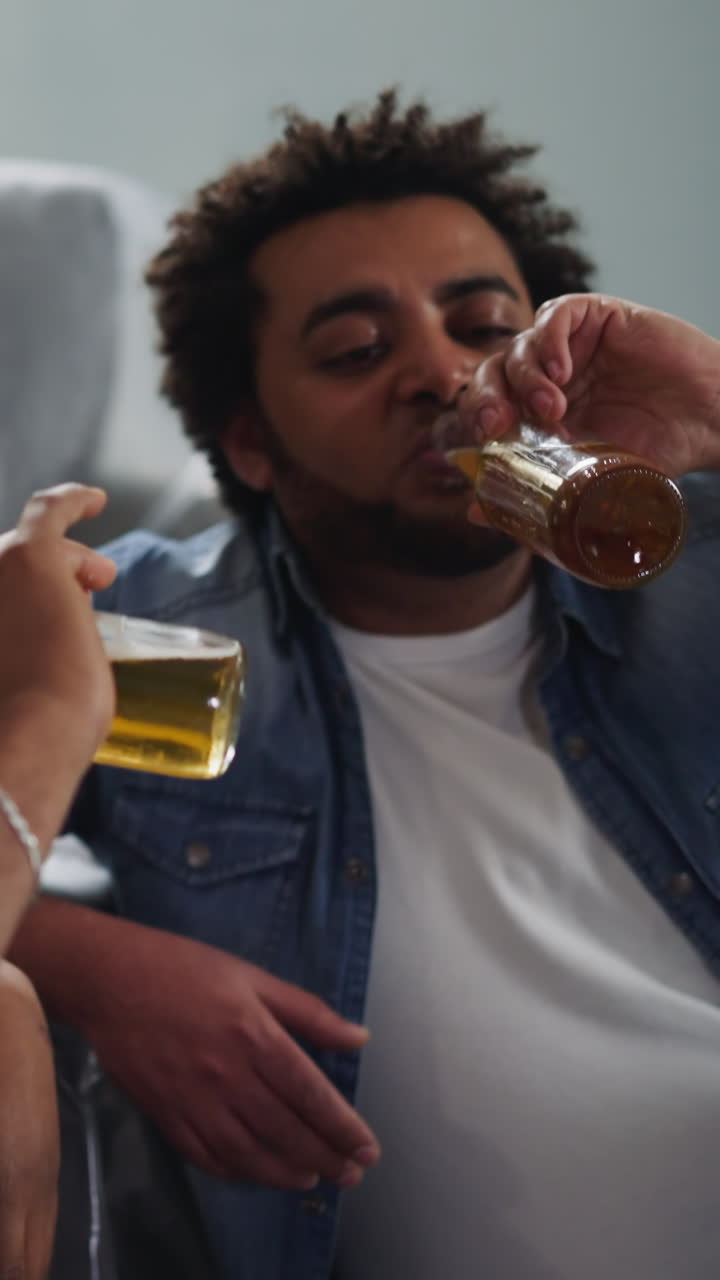 Positive black builders drink delicious beer sitting on floor near foiled sofa closeup. African-American guys relax after hard work upon restoration