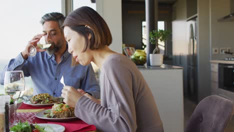 happy diverse couple sitting at table in dining room, eating dinner and drinking wine