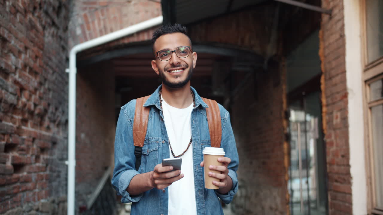 Smiling Young Man Walking in City Street with Smartphone and Coffee