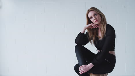 Fashion-Portrait-Of-Serious-Young-Woman-Sitting-Against-White-Studio-Wall