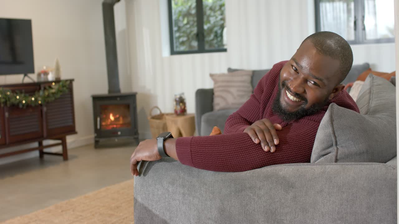 Noticing camera African American man leaning on gray sofa turning head and smiling in living room