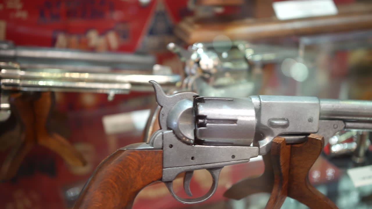 A vintage revolver displayed on a wooden stand inside a glass case, with blurred firearms and signage in the background of the museum or collection setting