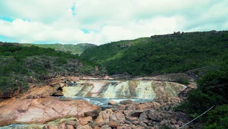 Tilting-up-shot-revealing-the-beautiful-Donana-waterfall-with-colorful-rocks,-dirty-water,-and-surrounded-by-green-tropical-hills-in-the-Chapada-Diamantina-National-Park-in-Bahia,-northeastern-Brazil