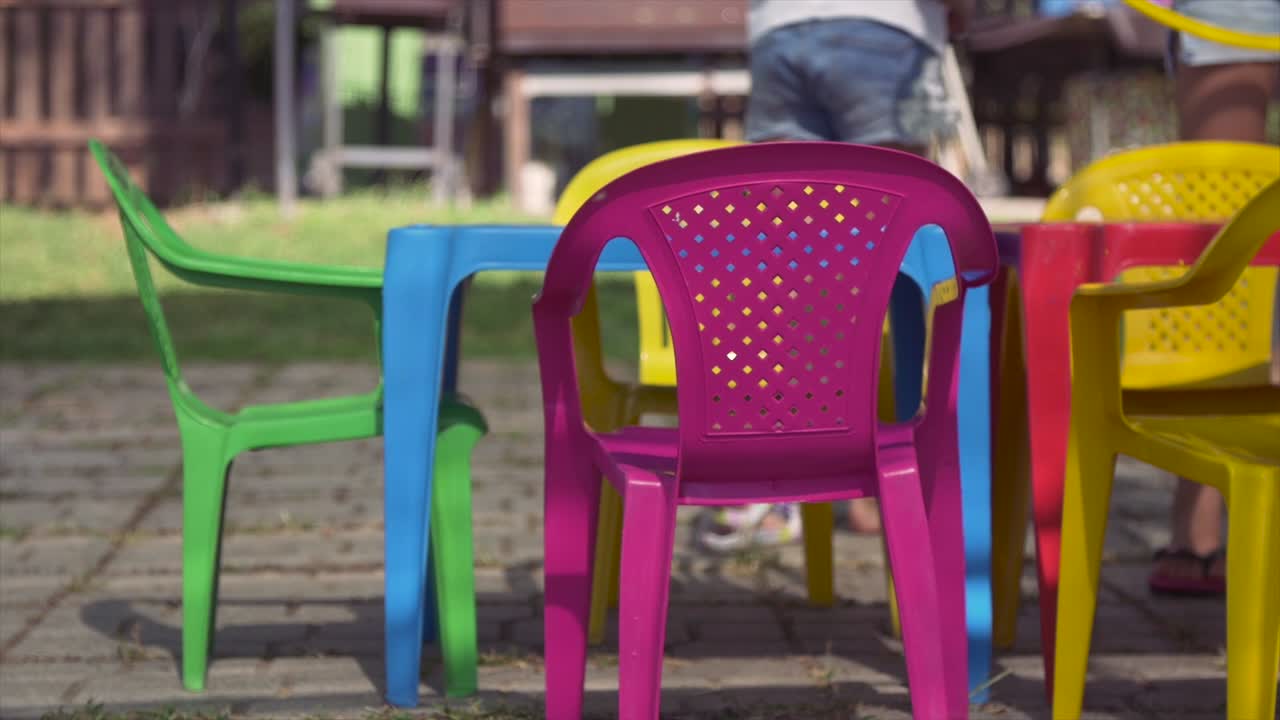 Jeunes enfants joyeux en camp d'été, assis sur des chaises en plastique colorées, buvant et mangeant des collations, s'amusant à l'extérieur dans un champ d'herbe verte, fête d'enfants au ralenti.