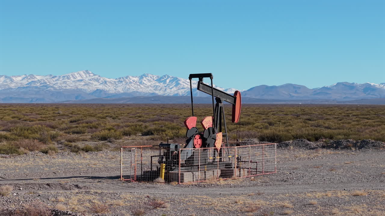 Drone shot orbiting over oil pumpjack in the dry scrublands of Mendoza, Argentina, with the Andes mountains rising in the background under clear blue skies