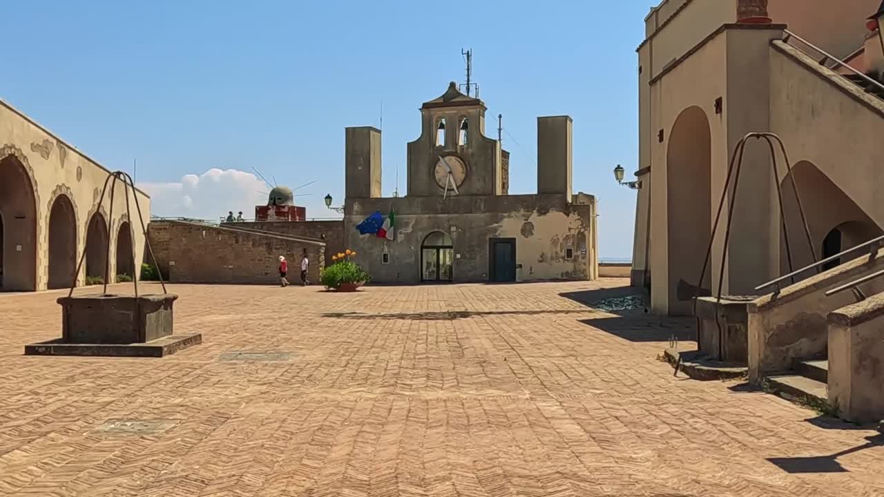 A tranquil courtyard featuring a historic clock tower under a clear blue sky, surrounded by arches and stone structures.