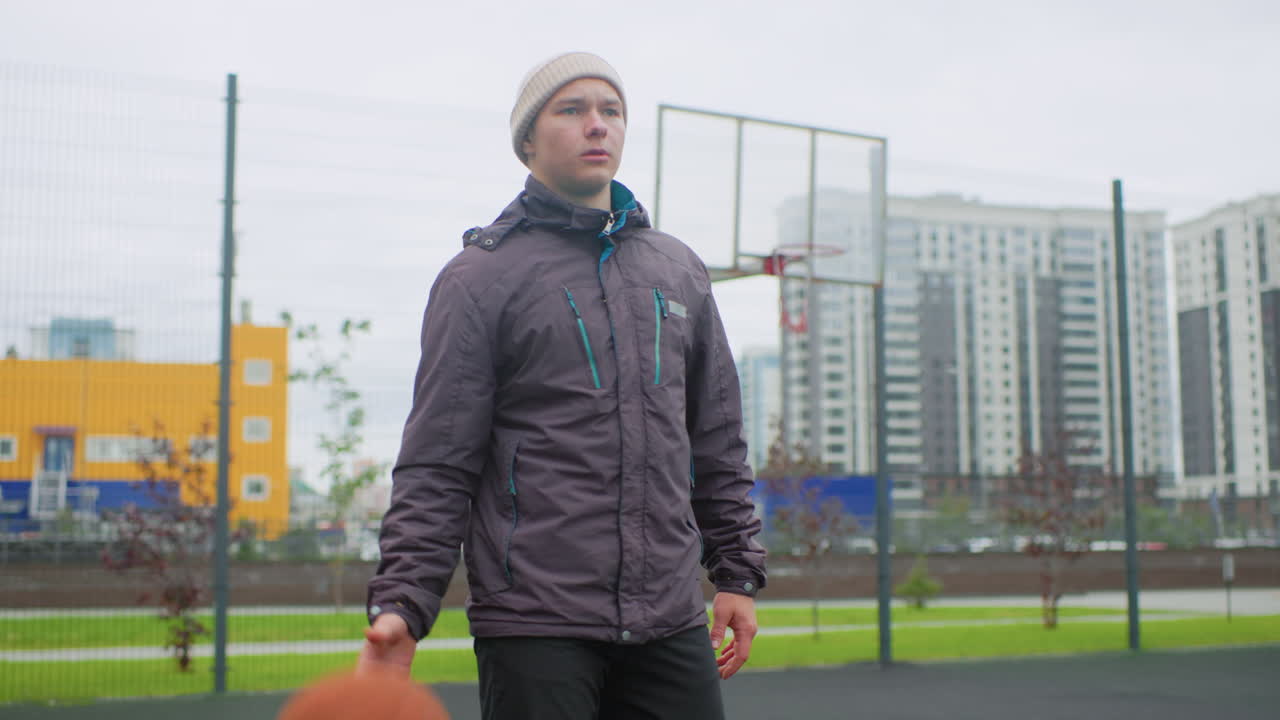 Young man dribbling basketball on court, wearing winter jacket and beanie, apartment towers and fenced hoop in background, overcast sky, cold mood, solo practice and walking shots, casual athlete