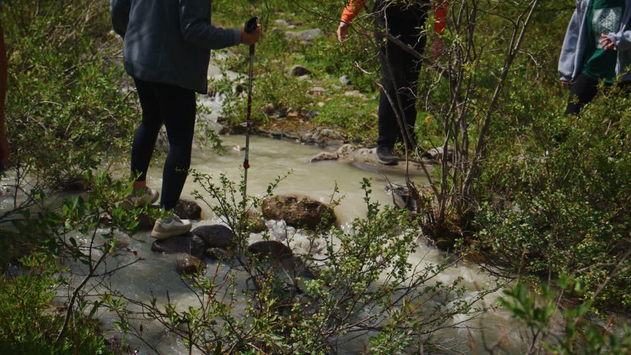 Hikers navigate shallow, rocky stream with trekking poles. Dense green bushes line water's edge. One hiker focuses intently on step, balancing carefully. Group moves slowly through wet terrain