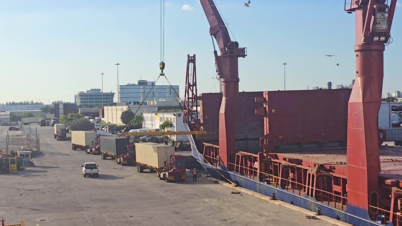 An elevated daytime view of a large geared cargo ship docked at a port terminal, with its crane hook lowered as yard trucks and trailers line up to load or unload shipping containers.
