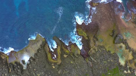 Beautiful-cinematic-shot-of-waves-crashing-on-the-rocky-coast-creating-a-foam-pattern-on-the-reef-view-from-a-drone-during-the-day