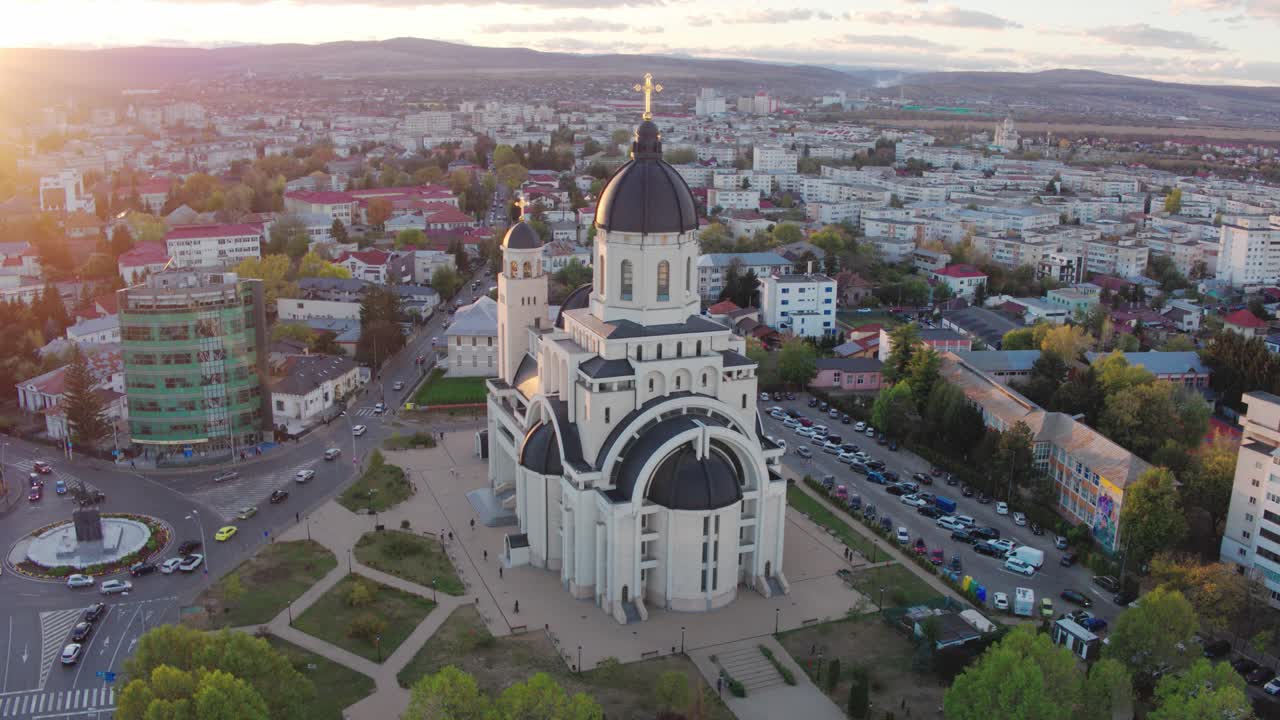 A cinematic aerial shot slowly pans right, showing a majestic Orthodox cathedral and the city of Targu Mures, Romania, bathed in the warm, glowing light of a golden hour sunset