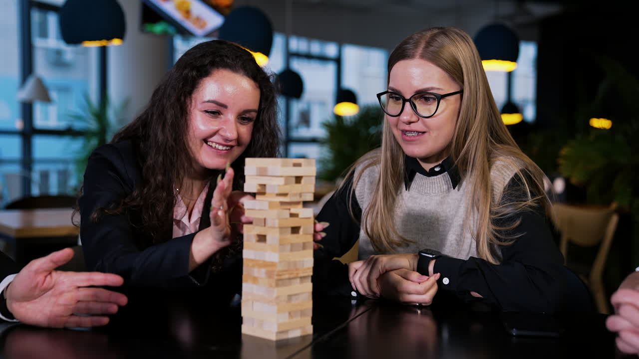 Colleagues having a break from work playing board games. Two ladies pull the wooden brick from a pile in jenga game.