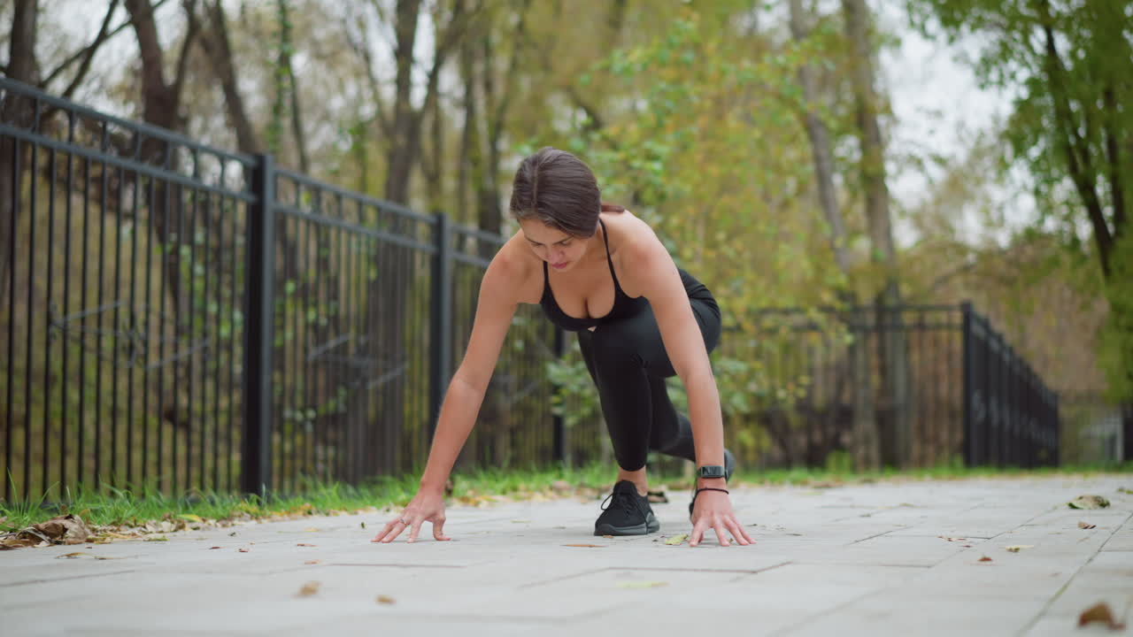 Sporty woman in black singlet and leggings preparing to run in outdoor park, crouched and ready to sprint, with trees and iron fence in background, focused on taking off and running