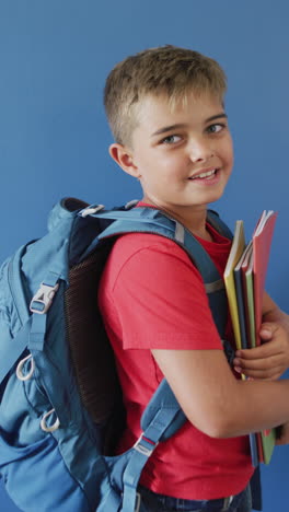 vertical video of happy caucasian boy holding books on blue background, copy space, slow motion