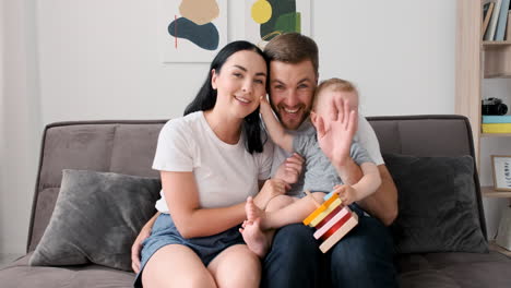 padres amorosos sentados en el sofá en la sala de estar con su adorable niño pequeño mientras miran la cámara durante una videollamada en casa