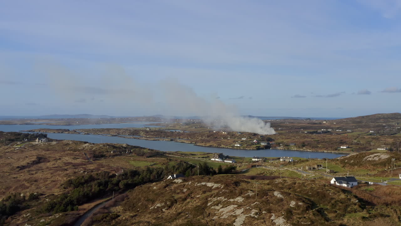 Wide aerial tracking shot over Sky Road coast, overlooking farmland where smoke drifts in the distance. Connemara landscape