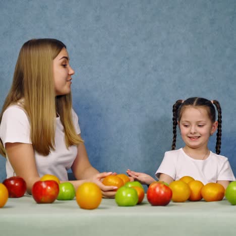 fresh organic fruit on the table. little girl together with mother playing with ripe fruits on blue background. woman and small girl in white t-shirts at home.