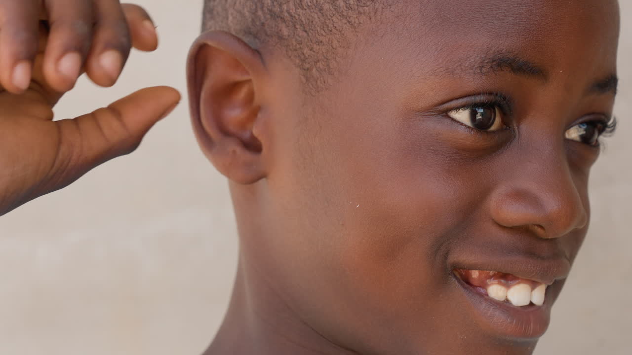 Primo piano di un ragazzo che si tocca l'orecchio, con un sorriso giocoso e una risata spontanea. La calda luce del sole esalta la texture della pelle. Capelli corti e uno sfondo rilassato di un muro di cortile o scuola, catturando un gesto giovanile e un momento spontaneo.