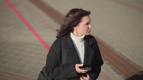 lady walking outdoors on an interlocked path, touching her hair, with phone in hand, looking around as she strolls through the urban landscape