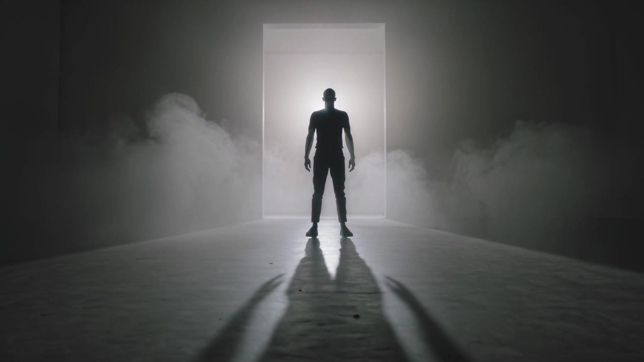 Uomo che avanza in abbigliamento scuro aderente verso una luce intensa, entrando in un portale sul palco, con nebbia che si diffonde.