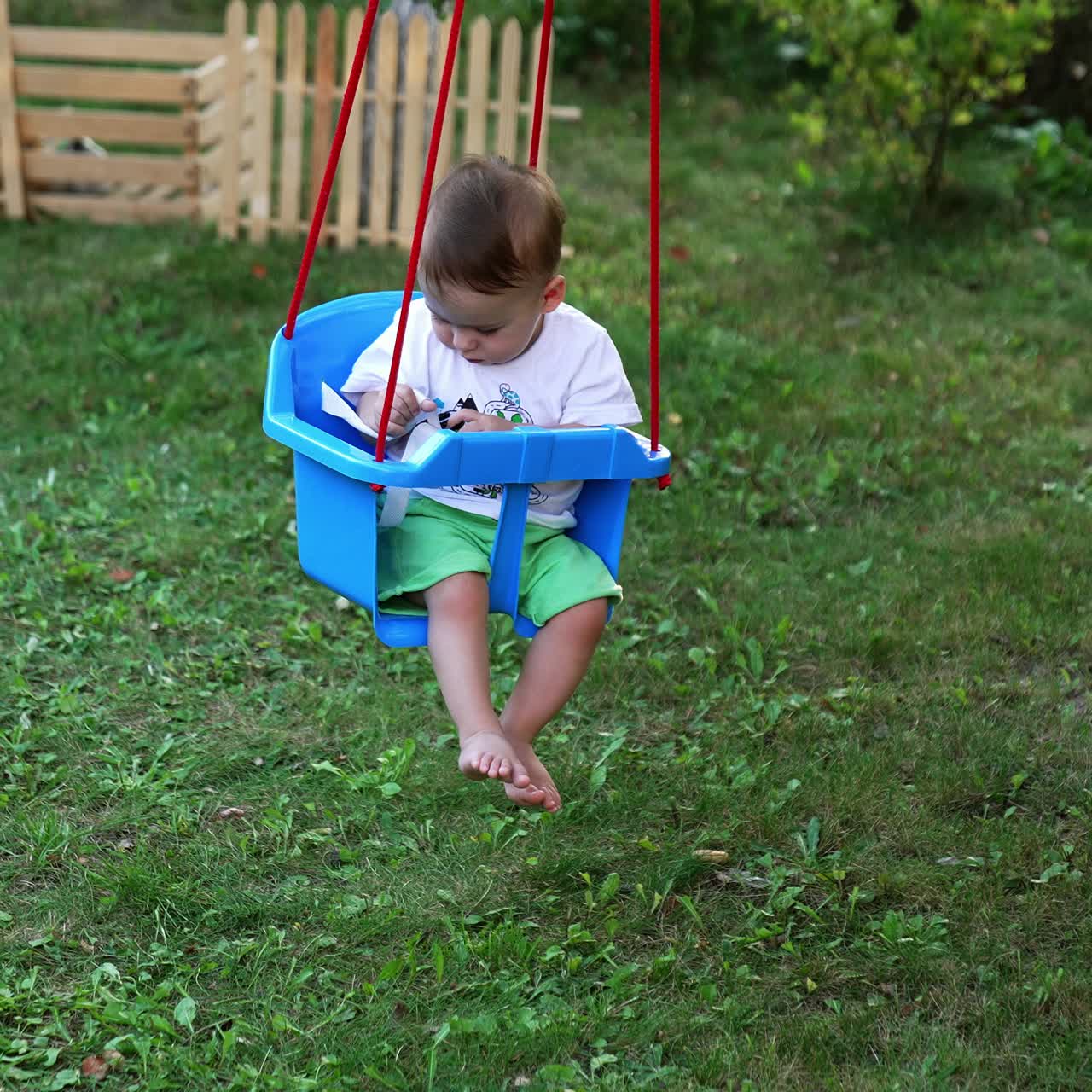 Little boy swaying in a swing looking attentively at the fastener in his hands. Cute kid in the summer garden. Nature backdrop