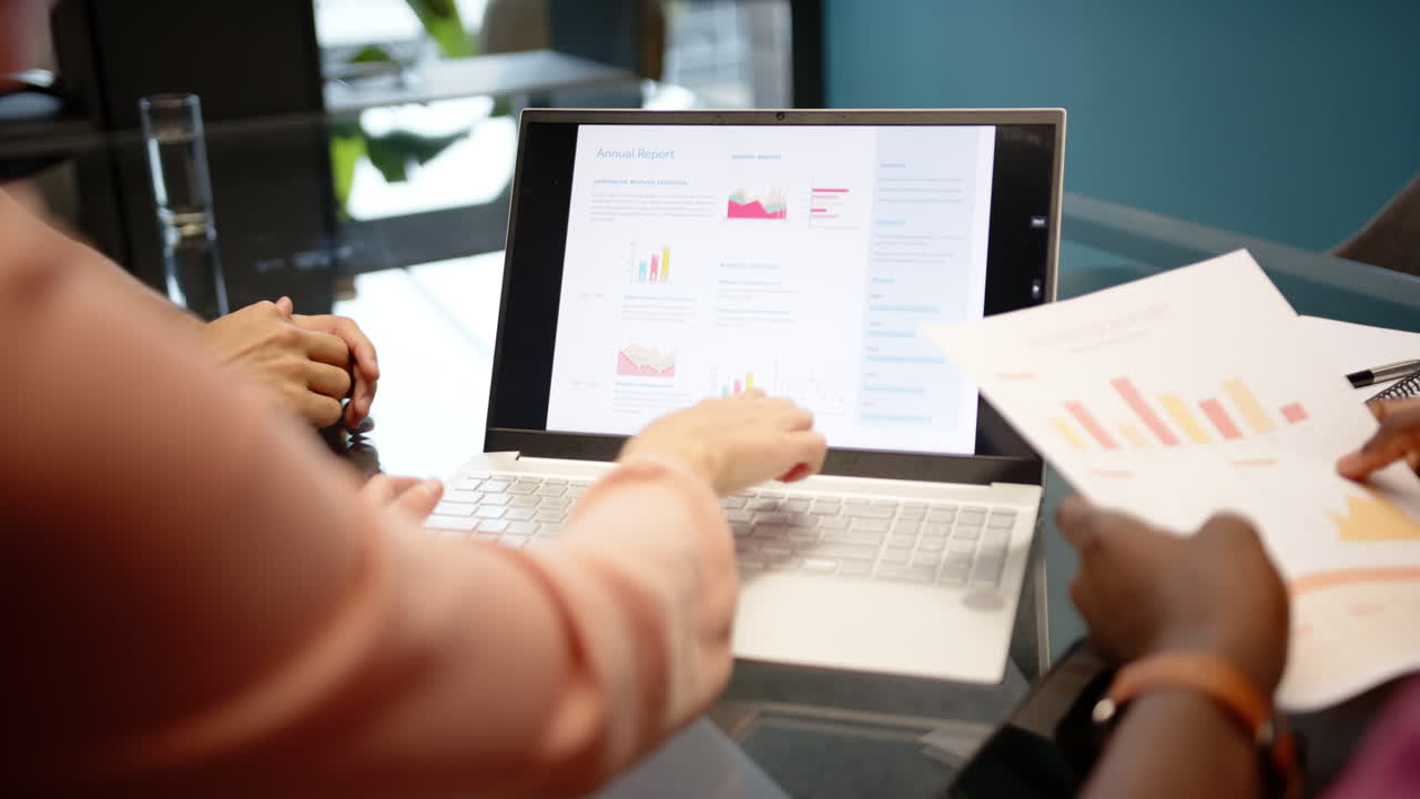 Pointing at laptop screen, diverse colleagues discussing annual report in office meeting