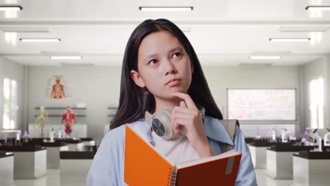 close up of asian teen girl student with a backpack reading book and thinking and looking around while standing in science laboratory