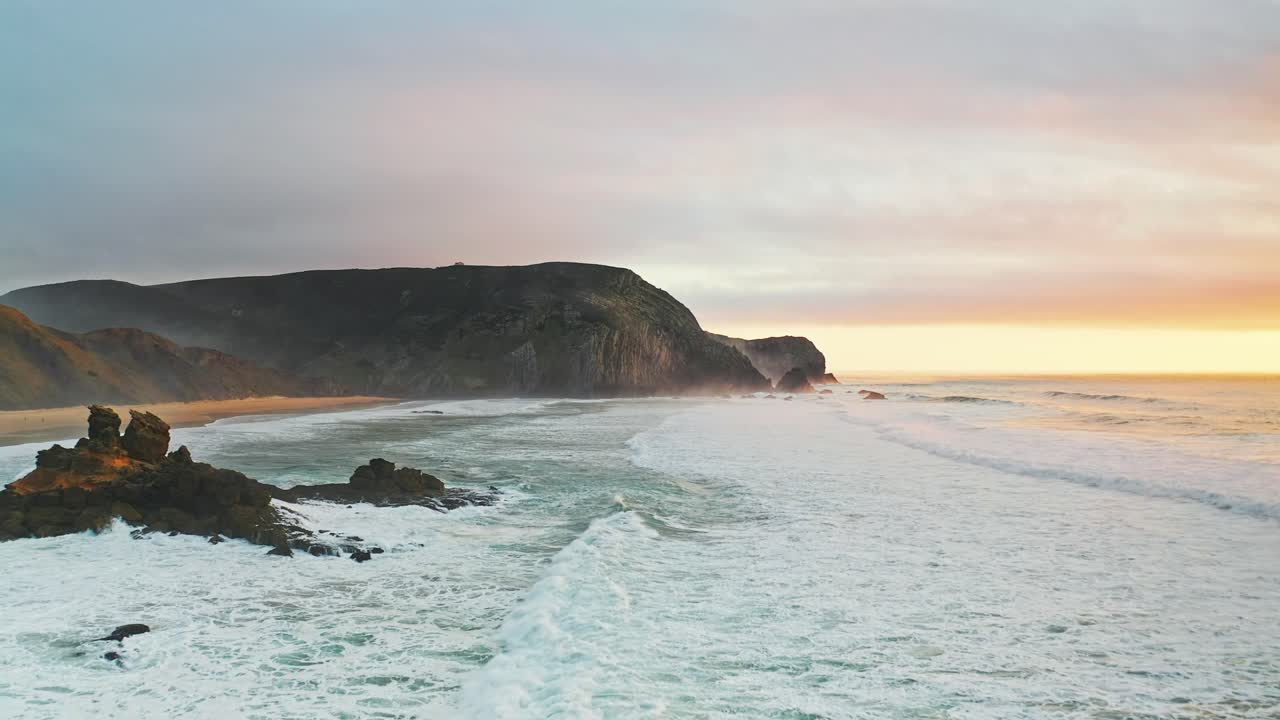Portugal beach cliffs on Atlantic ocean at sunset from aerial drone forwards shot with rough waves near rocky outcrop in water