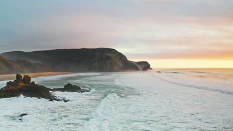 portugal beach cliffs on atlantic ocean at sunset from aerial drone forwards shot with rough waves near rocky outcrop in water