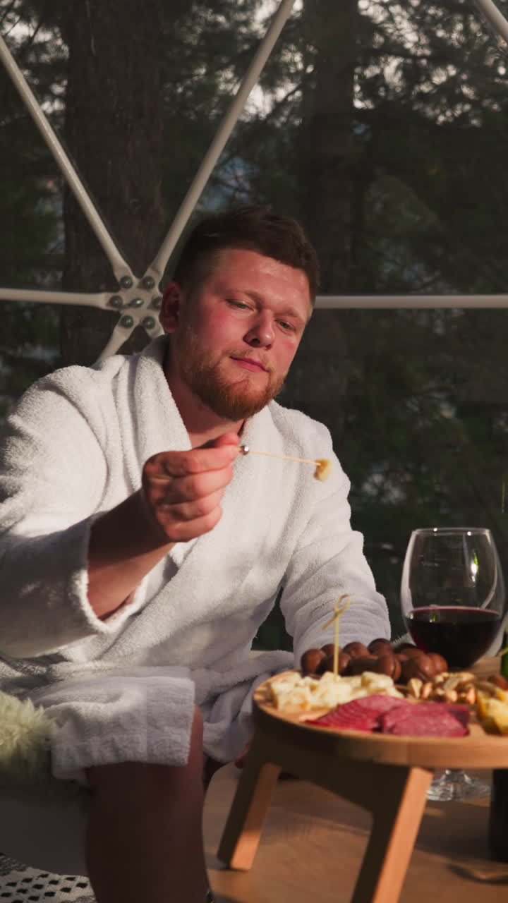 Man eats cheese on skewer at glamping round house. Relaxed guest in soft bathrobe enjoys delicacy snacks sitting by panoramic window. Luxury vacation