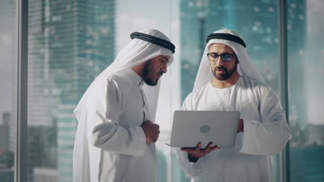 dos hombres de negocios musulmanes exitosos con trajes tradicionales blancos de pie en una reunión de oficina, negociando y hablando sobre oportunidades financieras, usando una computadora portátil.