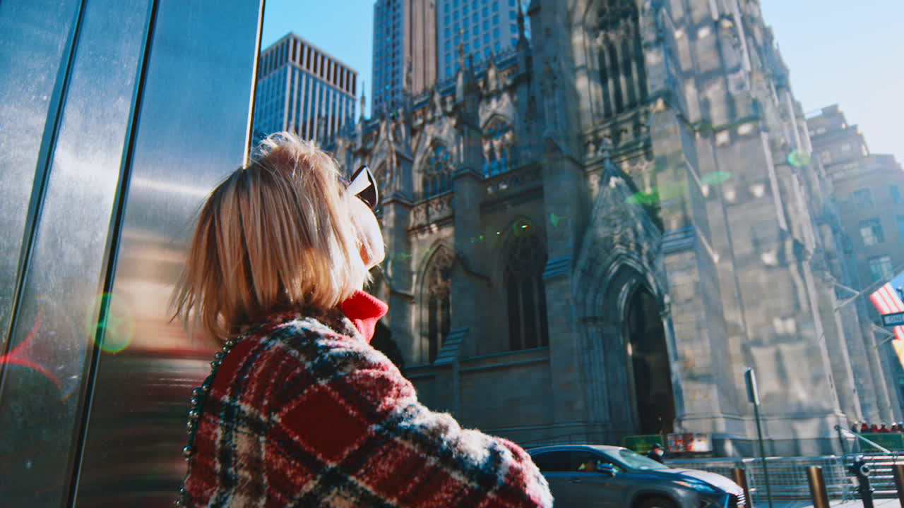 New York, USA, 2 aprile 2026: Donna in cappotto a quadri che guarda la cattedrale gotica a Manhattan. Vista laterale di una donna bionda con occhiali da sole e cappotto a quadri in piedi davanti a una chiesa in pietra sotto il sole splendente.