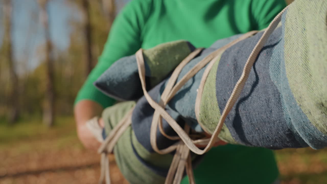 Vrouw bindt kampeerdeken vast voor het inpakken, draagt een groene trui, handen lussen koord om opgerolde stof, bevestigen knoop voor transport, close-up shots van grip en beweging, herfstbos op de achtergrond, efficiënt