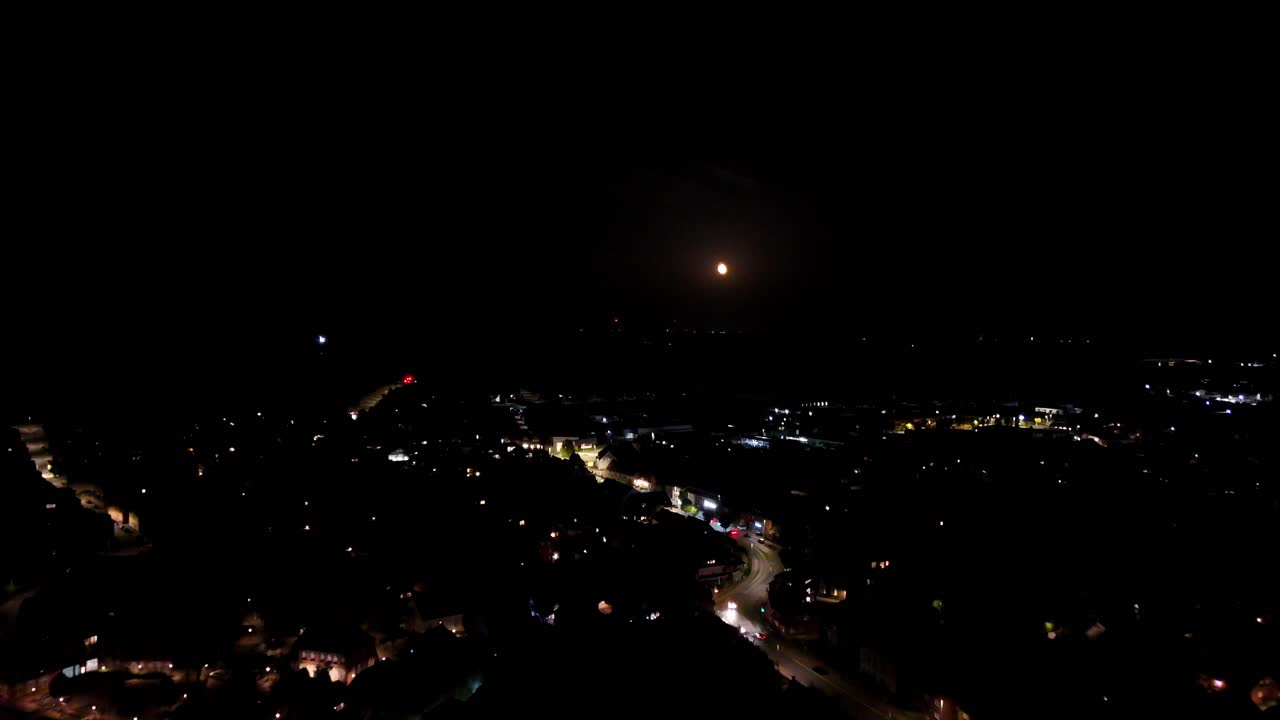 Slow drone flight over illuminated Main Street with driving cars at night sky. Aerial forward wide shot. Golden full moon at dark sky. Panorama view. Houses and homes in American city
