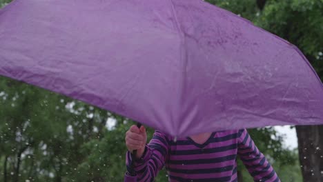 nahaufnahme eines jungen kaukasischen mädchens in gestreiftem hemd mit lila regenschirm, das draußen auf und ab springt und wasser in einer pfütze spritzt, statisches porträt in zeitlupe