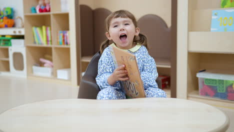 Fröhliches-Lächelndes-Mädchen,-Das-Mit-Einem-Buch-In-Der-Bibliothek-Spielt---Früherziehungskonzept-Für-Kinder