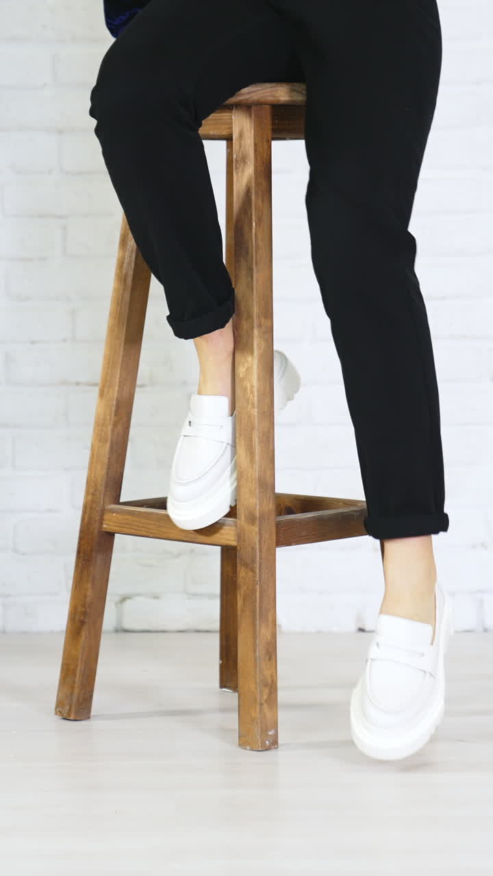 Girl model climbs up on the high wooden stool and puts her shoes on the footrest. White new modern footwear demonstration. White wall background. Vertical video