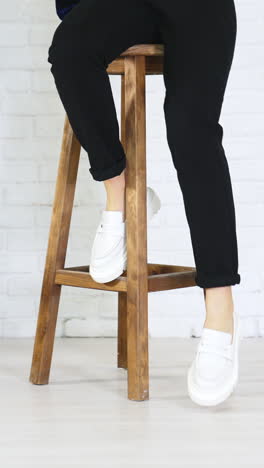 girl model climbs up on the high wooden stool and puts her shoes on the footrest. white new modern footwear demonstration. white wall background. vertical video