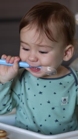 one-year old toddler sitting at the feeding table with spoon in mouth. child turns to bagels left in front of him. blurred background. vertical video