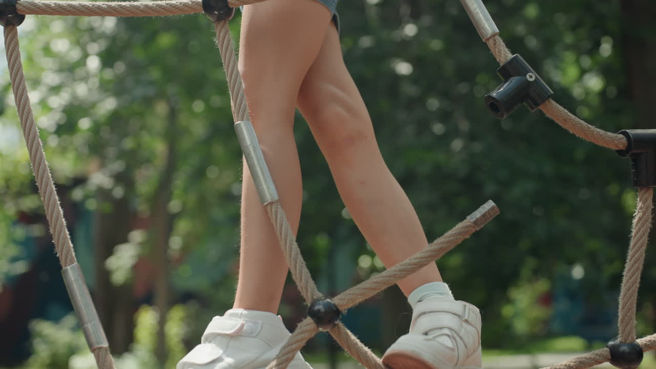 Giovane figlia che si arrampica su una rete di corda in un parco, mani ferme che afferrano la corda ruvida, scarpe da ginnastica bianche che calpestano tra i nodi, alberi illuminati dal sole e sfondo con bokeh, avventura estiva giocosa e fiduciosa.