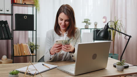 Happy-rich-business-woman-counting-money-cash-dividends-using-smartphone-calculator-in-home-office