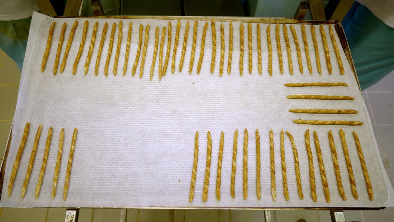 Hands arranging grissini breadsticks on a baking tray with parchment paper in a bakery kitchen, artisan dough sticks lined up for baking, snack production and food preparation