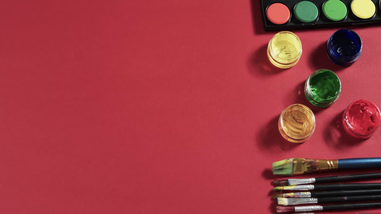 vista aérea de pinturas y pinceles dispuestos sobre un fondo rojo con espacio de copia, en cámara lenta
