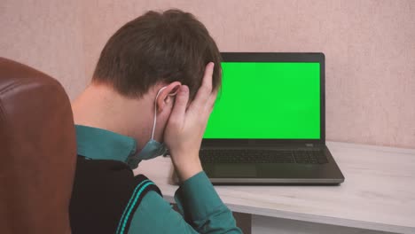 un tipo con una máscara de protección emocionalmente enojado por una conexión remota en una videoconferencia y golpea sus puños en una computadora portátil, pantalla verde