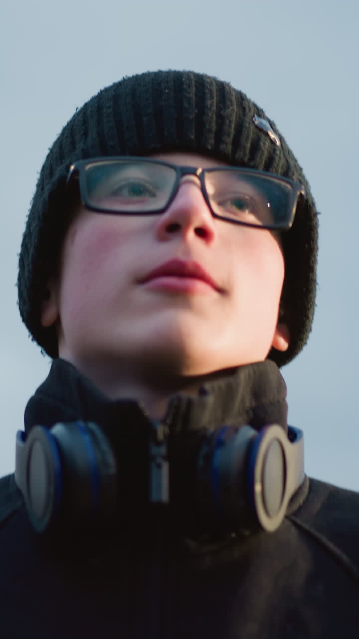 A close-up of a young boy wearing glasses and a black outfit with a headset around his neck, with a blur view of tree in the background