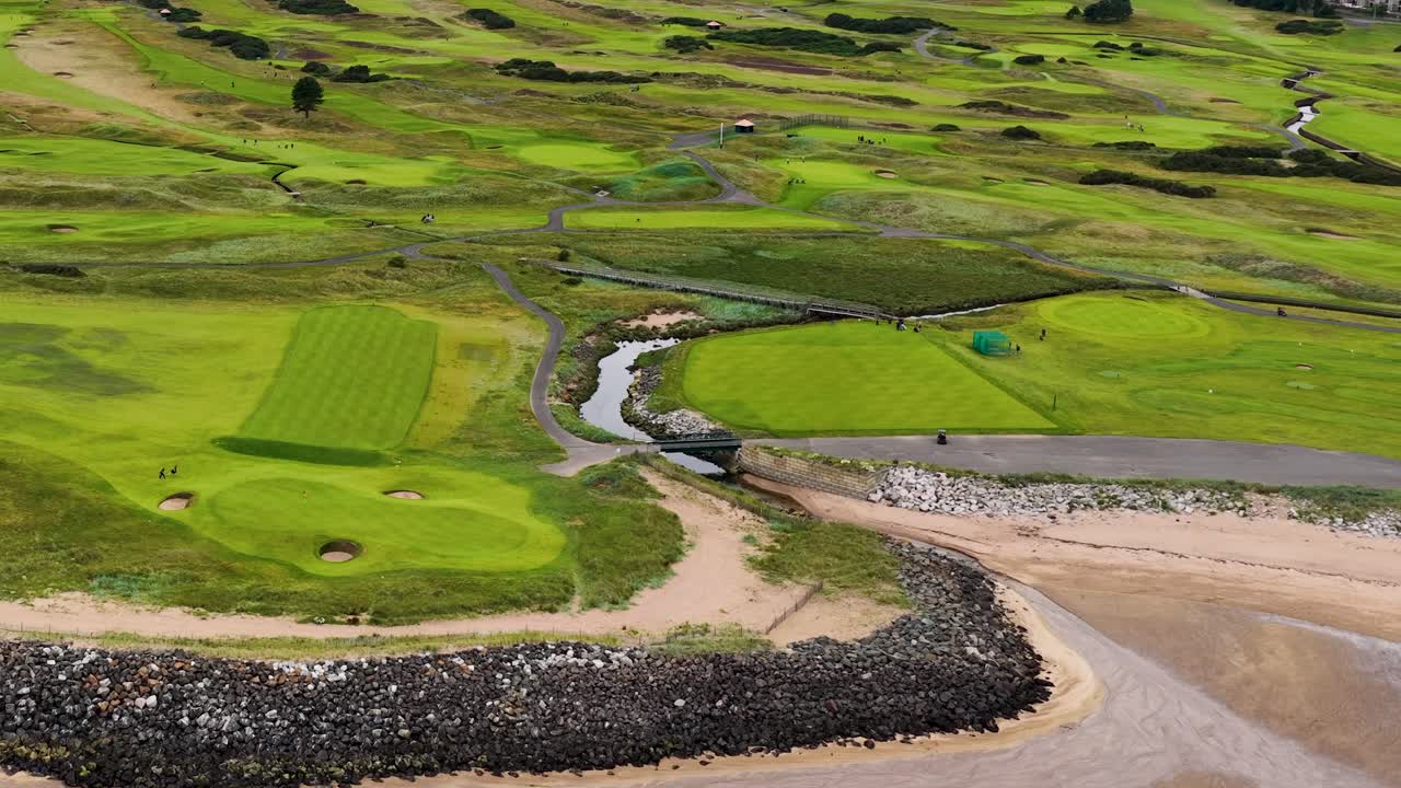 Drone pans above lush golf course, sandy beach, rolling fairways, and coastal landscape in daylight