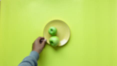 a hand reaches for a green apple on a yellow plate