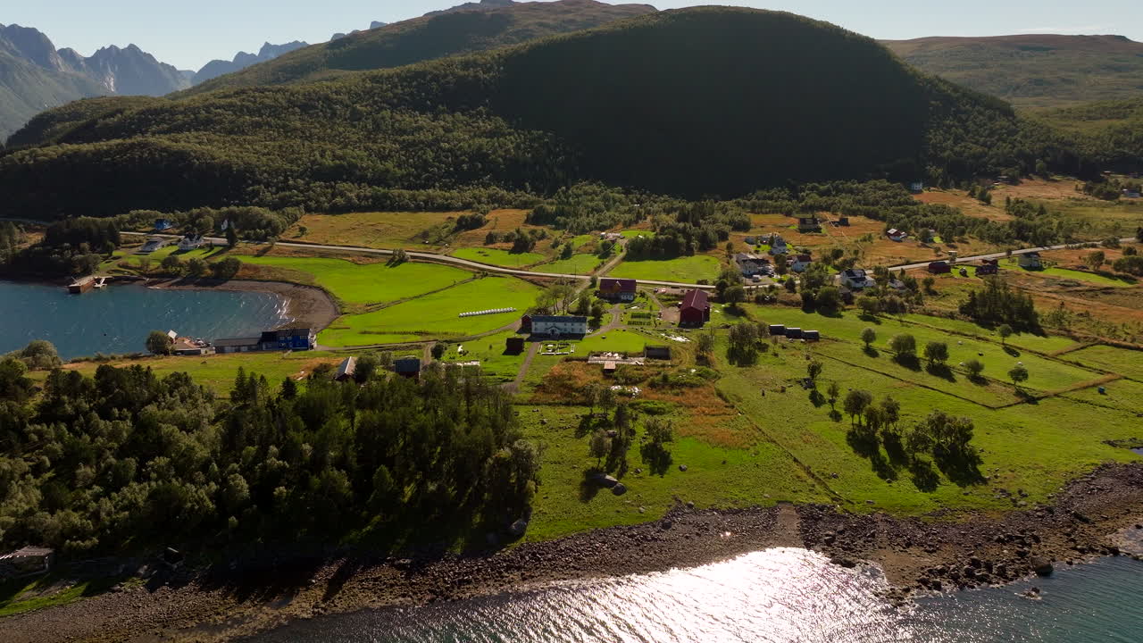 Kvitnes Gård, Hadsel Municipality, Norway - A Peaceful Coastal Farm Sits Beside Clear Waters, Surrounded by Wide Green Fields and Mountain Landscapes - Drone Flying Backward