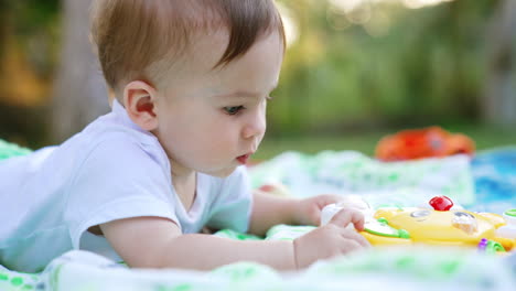 lovely caucasian baby busy with a bright toy. calm little child lying on the blanket on the ground outdoors. close up.