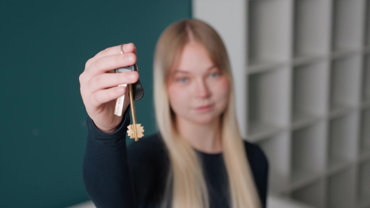 Mujer caucásica entregando llaves de un apartamento, retrato tranquilo en primer plano en una sala de estar moderna y vacía, propietaria rubia extendiendo la mano con llavero de latón, fondo de estanterías minimalistas, hito esperanzador.